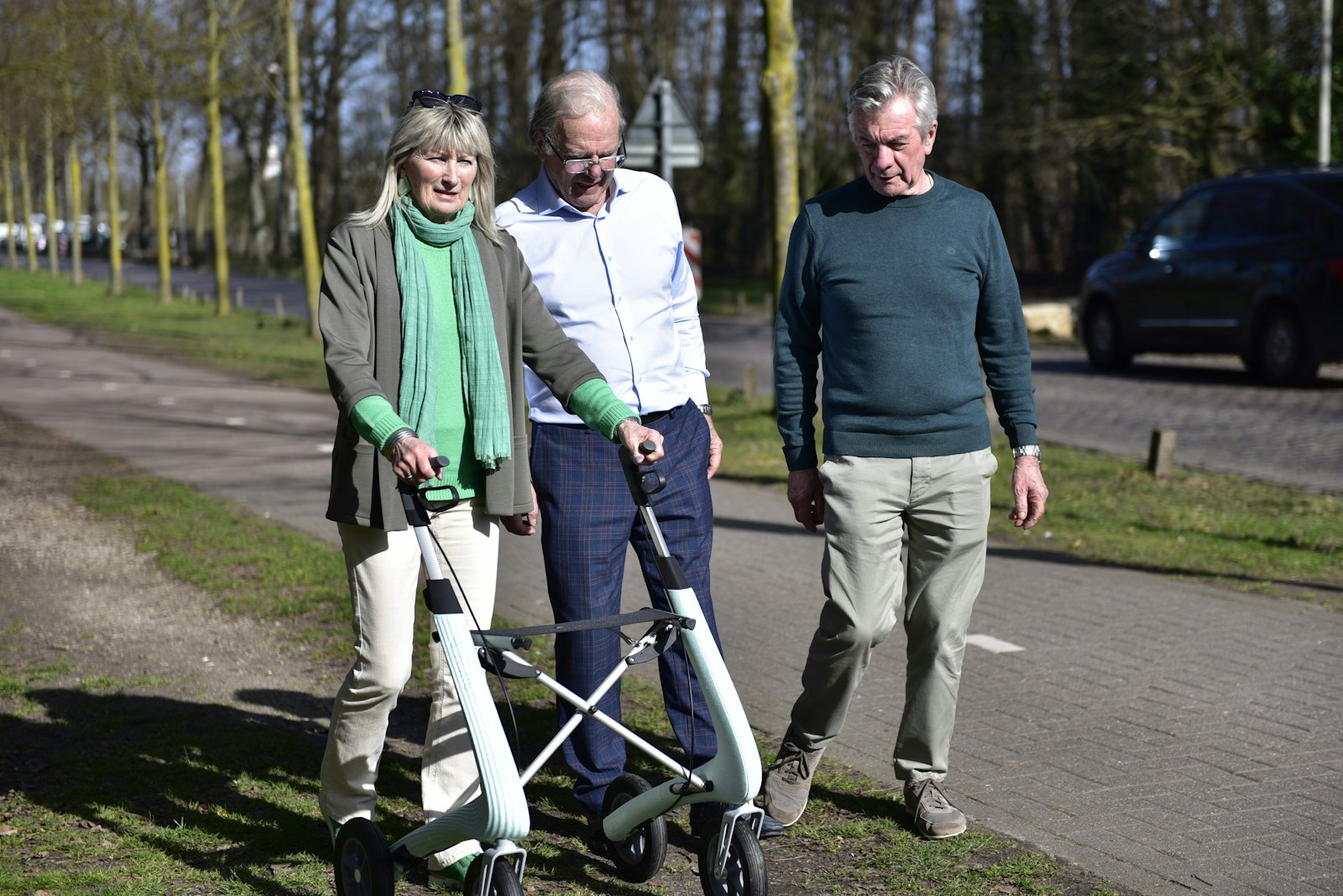 An older woman walks outdoors with a rollator, with 2 people beside her.